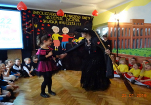 Na scenie na tle dekoracji z okazji Dnia Babci i Dziadka stoi Latająca Babcia w przebraniu czarownicy z miotłą oraz dziewczynka przebrana za czarownicę.